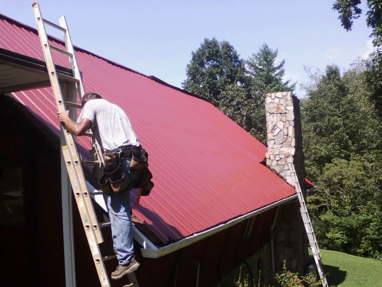 Roofer on a ladder beside a red metal roof and stone chimney — Soffit & Fascia Repair by Roofing Brothers in Chepachet
